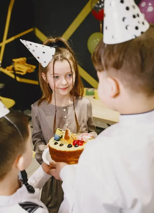 Girl in a party hat holding a fruit cake, surrounded by friends