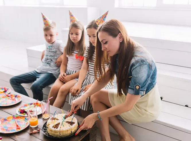 Mother cutting the cake for children in party hats, sitting on a bench